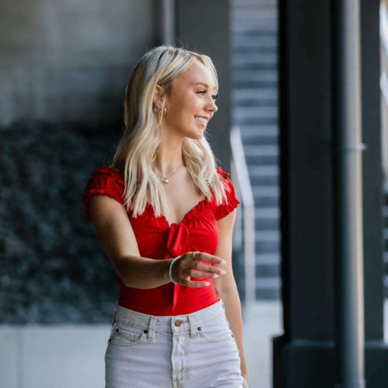 A blonde woman in a red top and white shorts walks in the foreground with a blurred urban background.