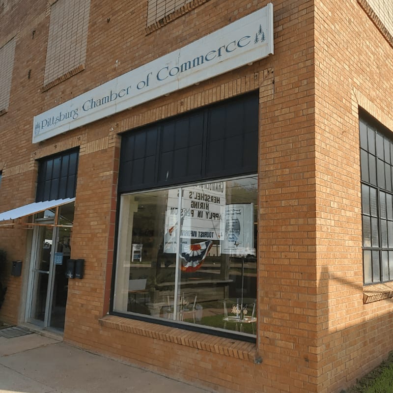 A brick building with a "Pittsburg Chamber of Commerce" sign above a large storefront window displays posters, while a smaller window and entrance are visible to the left.