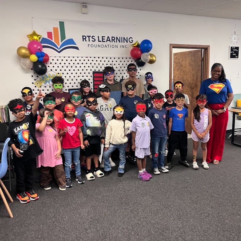 A group of children dressed as superheroes poses in front of a banner that reads "RTS LEARNING FOUNDATION INC."