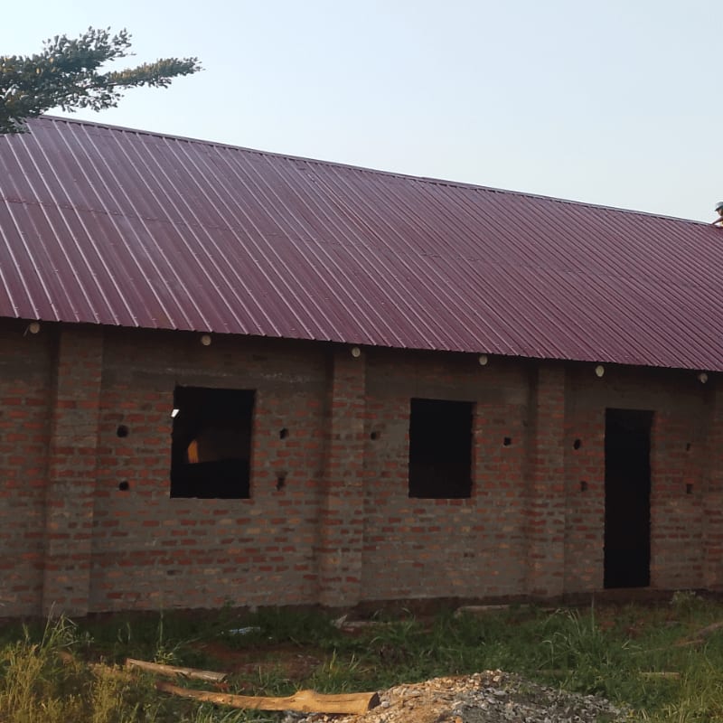 A brick building with a maroon metal roof stands unfinished in the foreground, with a clear sky and a few tree branches visible in the background.