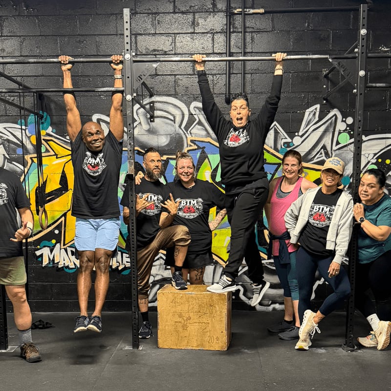 A group of people pose in front of graffiti-covered walls in a gym, with two individuals hanging from a pull-up bar in the foreground.