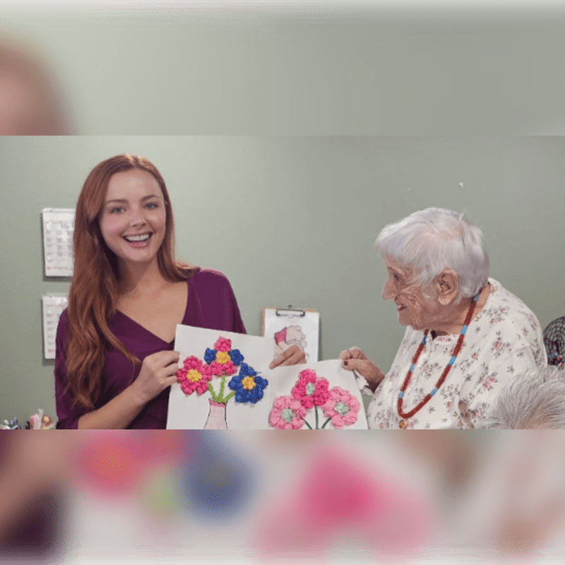 A young woman with red hair and an elderly woman with white hair smile while holding up colorful flower crafts against a pale green wall.