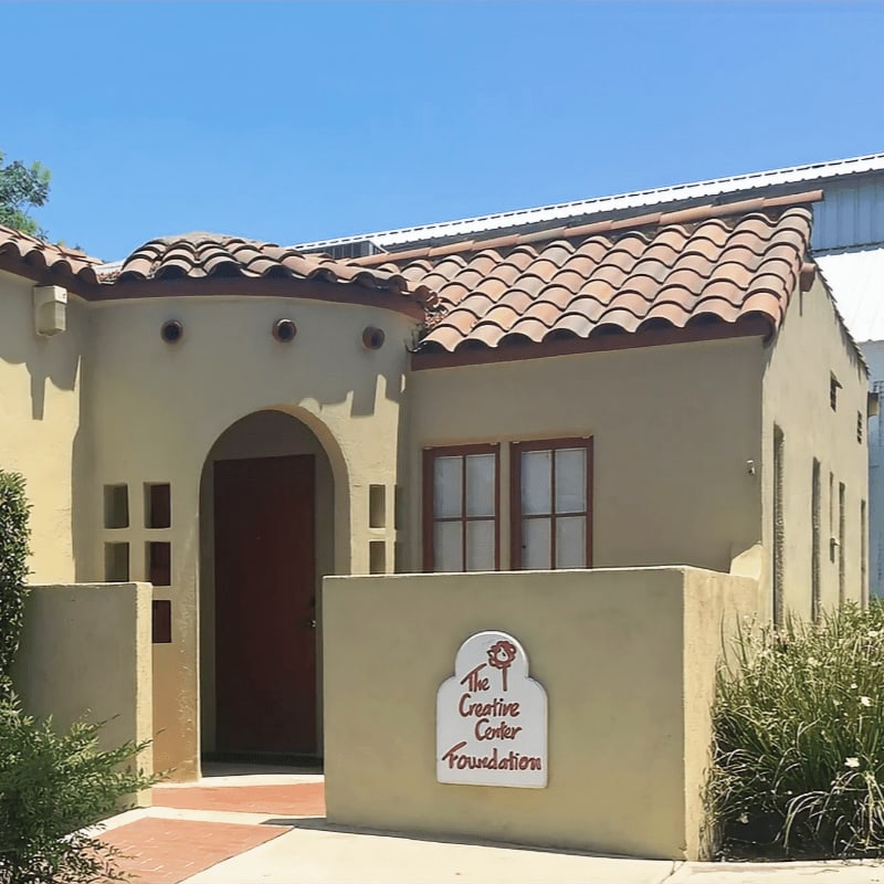 A stucco building with a red tile roof and a sign for "The Creative Center Foundation" in the foreground stands against a clear blue sky.