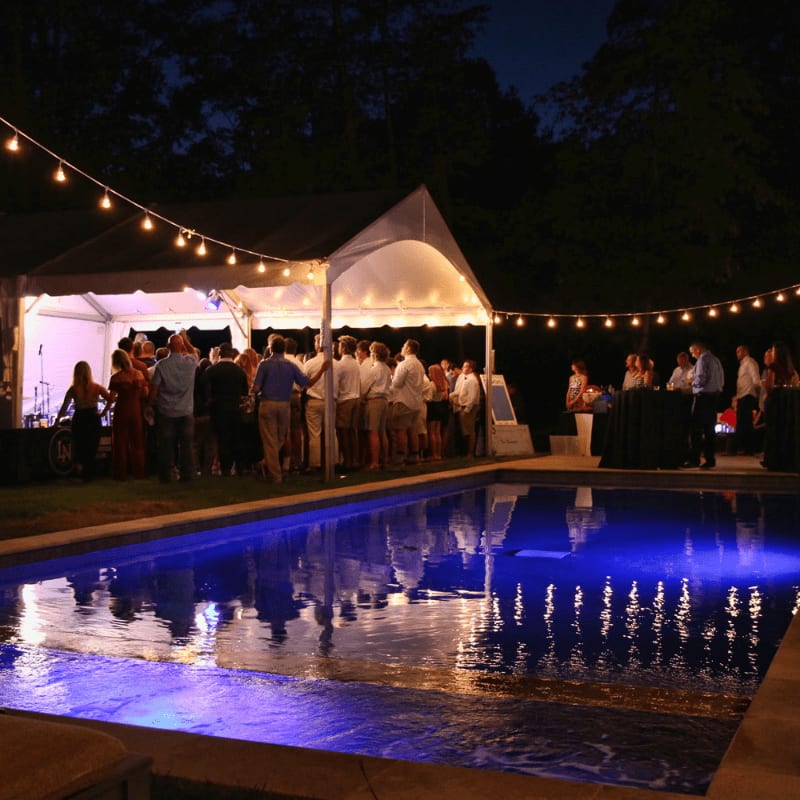 A brightly lit swimming pool with blue underwater lights reflects a lively outdoor party with guests mingling under string lights and a tent in the background.