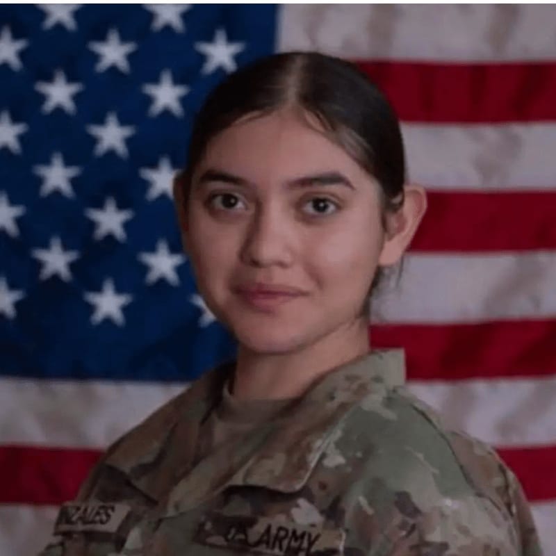A young woman in a US Army uniform stands in front of a blurred American flag.