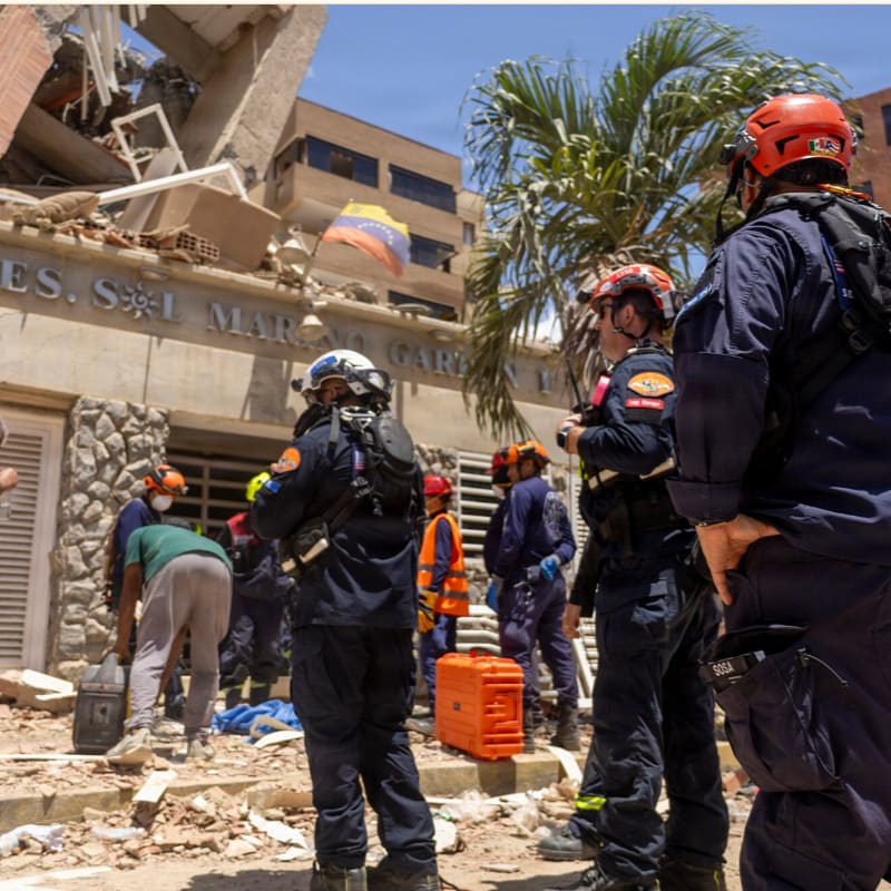Rescuers in helmets and tactical gear stand amidst rubble in the foreground, with a partially collapsed building and a Colombian flag visible in the background.