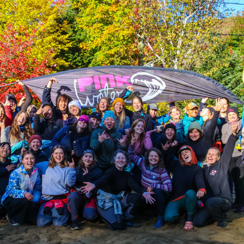 A group of women in casual clothing pose for a photo under a banner with colorful autumn trees in the background.