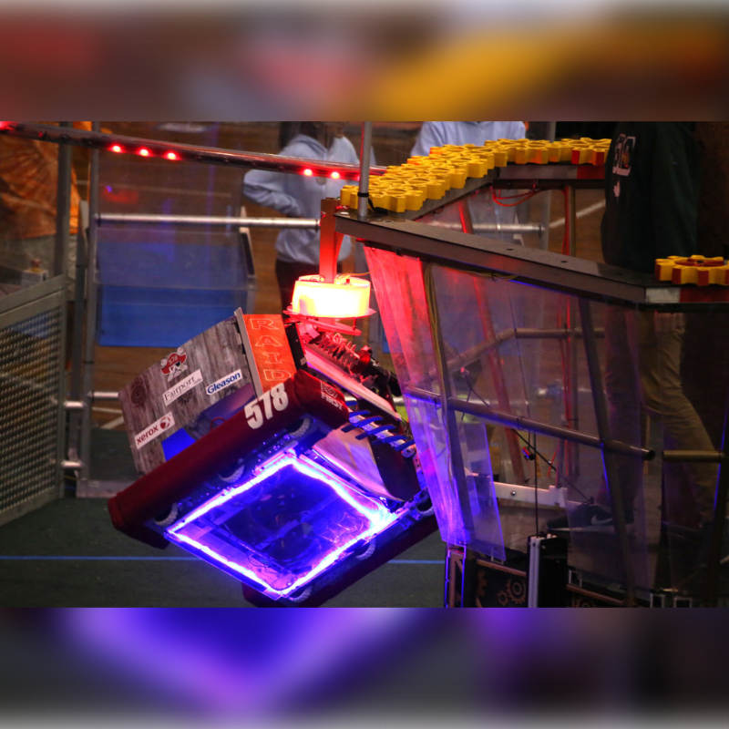 A robot with blue lights on its base is tilted on a ramp, with people and yellow game pieces in the background.