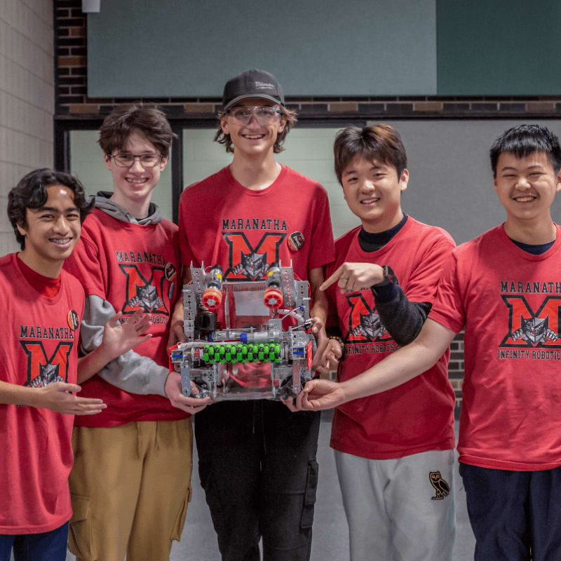 Five smiling teenagers in red shirts stand in the foreground holding a complex robot, with a brick wall and hallway visible in the background.