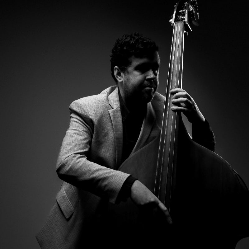 A man in a suit plays a double bass against a dark, moody background.