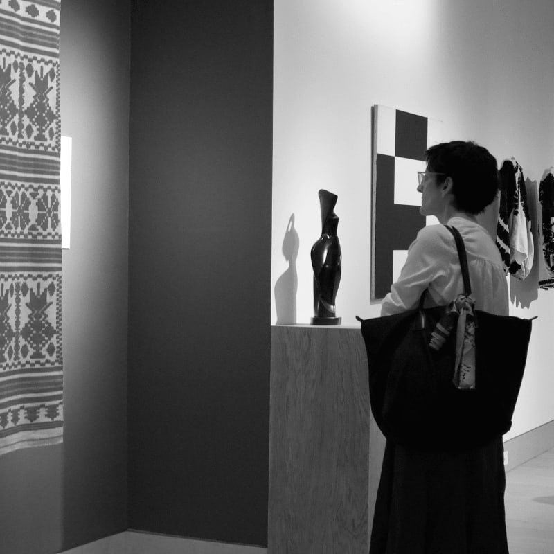 A woman with a large bag observes artwork, including a textile on the left and abstract paintings and sculptures on the right, in a gallery setting.
