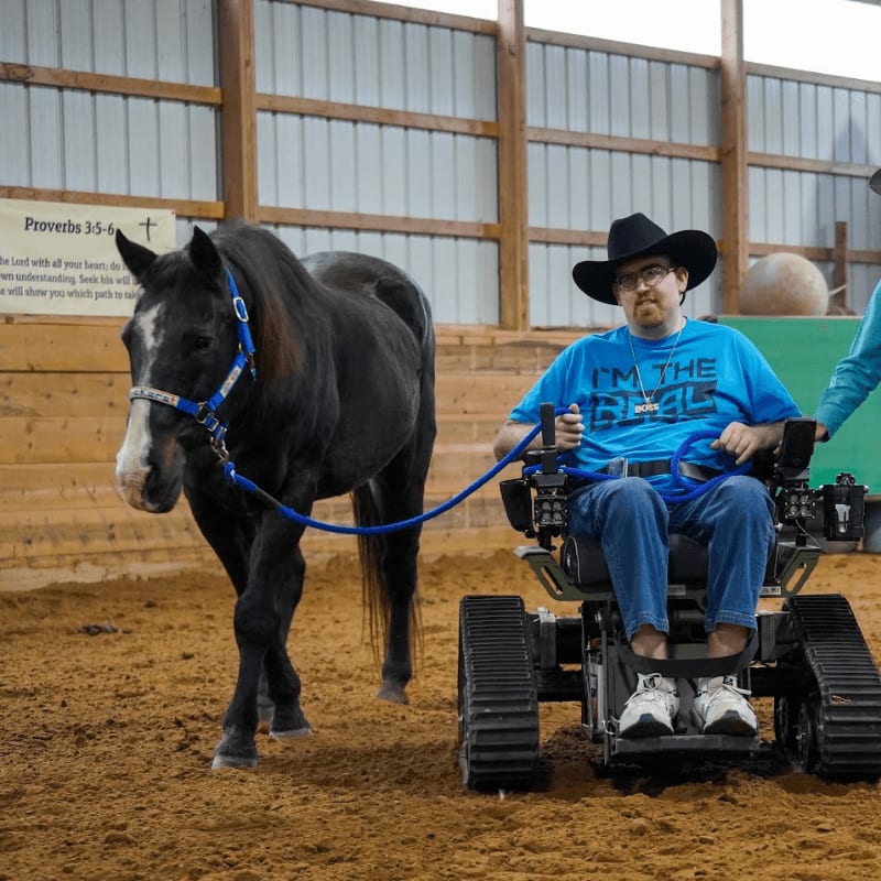 A man in a cowboy hat and blue t-shirt rides a tracked wheelchair beside a black horse in an indoor arena with a Bible verse on the wall.