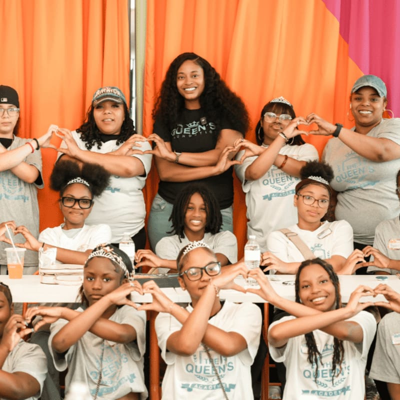 A group of young women and girls in matching t-shirts and tiaras form heart shapes with their hands in the foreground, against a backdrop of orange and pink fabric.