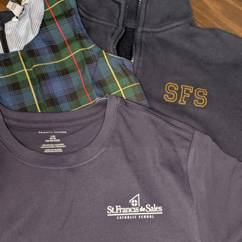 A dark grey t-shirt with "St. Francis de Sales Catholic School" printed on it lies in the foreground, with a plaid shirt and a navy blue quarter-zip sweatshirt with "SFS" embroidered on it in the background.