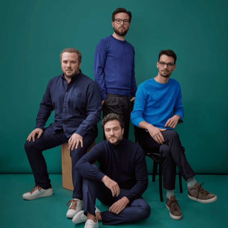 Four men in blue sweaters and shirts pose against a teal background, with one man sitting on the floor in the foreground.