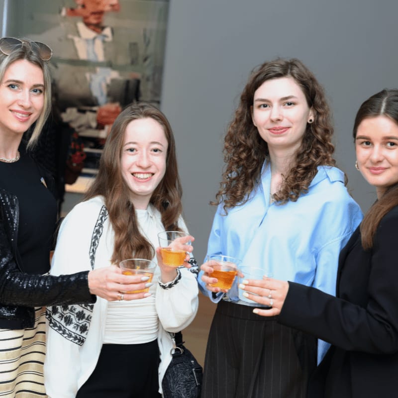 Four women smile and hold drinks in the foreground, with a blurred artwork in the background.
