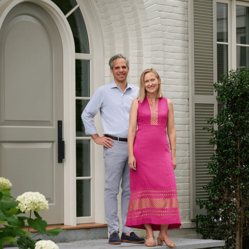 A couple stands on the steps of a white brick house with a large arched doorway, framed by white hydrangeas in the foreground.