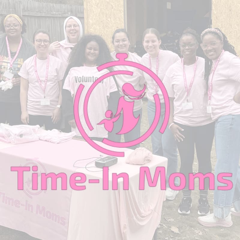 A group of women in pink shirts stand behind a table with a pink tablecloth in the foreground, while a blurred background shows a wooden structure and some outdoor elements.