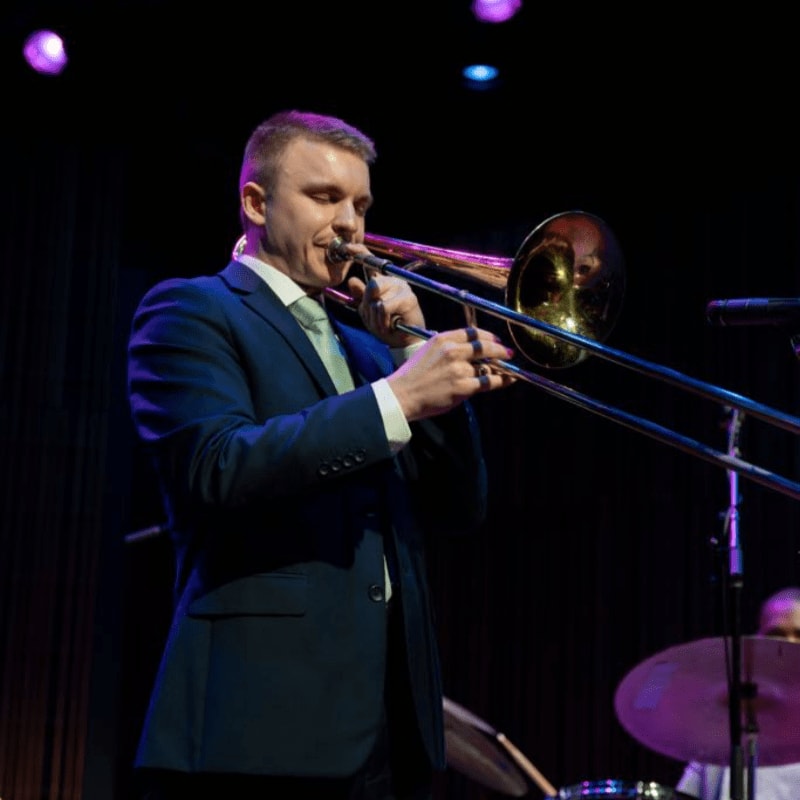 A man in a suit plays a trombone in the foreground, while a drummer is visible in the dimly lit background.