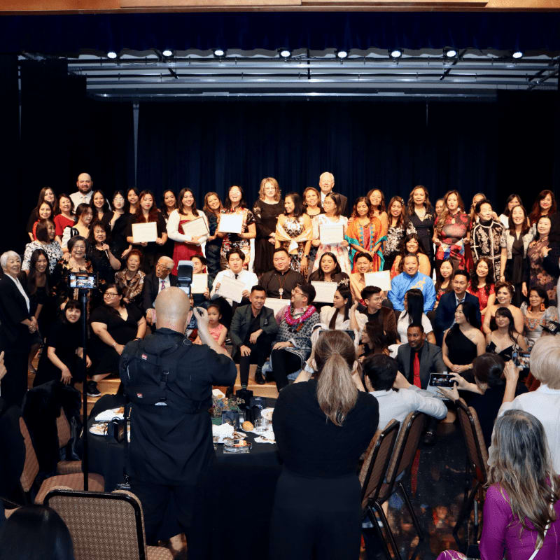 A large group of people in formal attire are posing for a photo on a stage with a dark backdrop, while a few individuals in the foreground are seated and looking towards the stage.