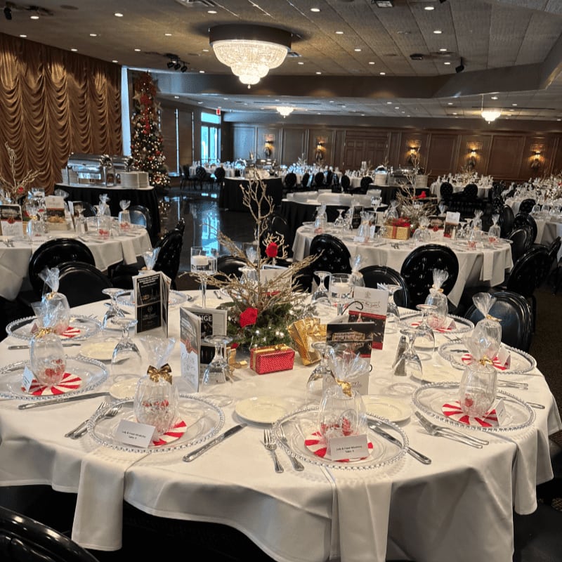 A festive banquet hall is set with round tables adorned with Christmas decorations and gifts in the foreground, while the background reveals more tables, a buffet, and a decorated Christmas tree.