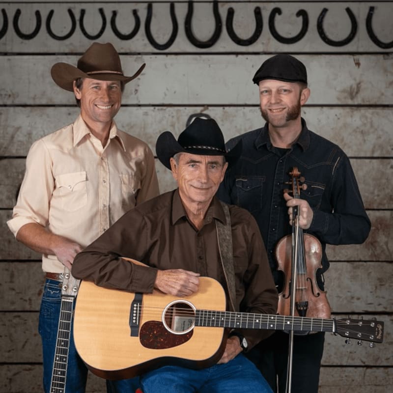 Three men in cowboy attire pose with musical instruments in front of a rustic wooden backdrop adorned with horseshoes.