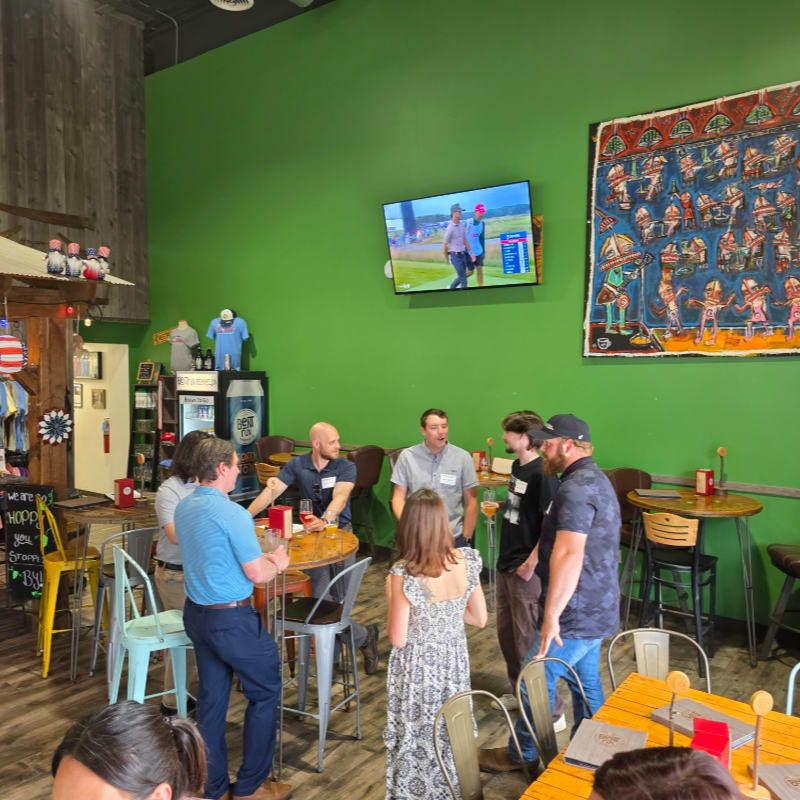 People gather in a brightly lit room with green walls, a television displaying a golf game, and a colorful tapestry, while others are seated at tables in the foreground.