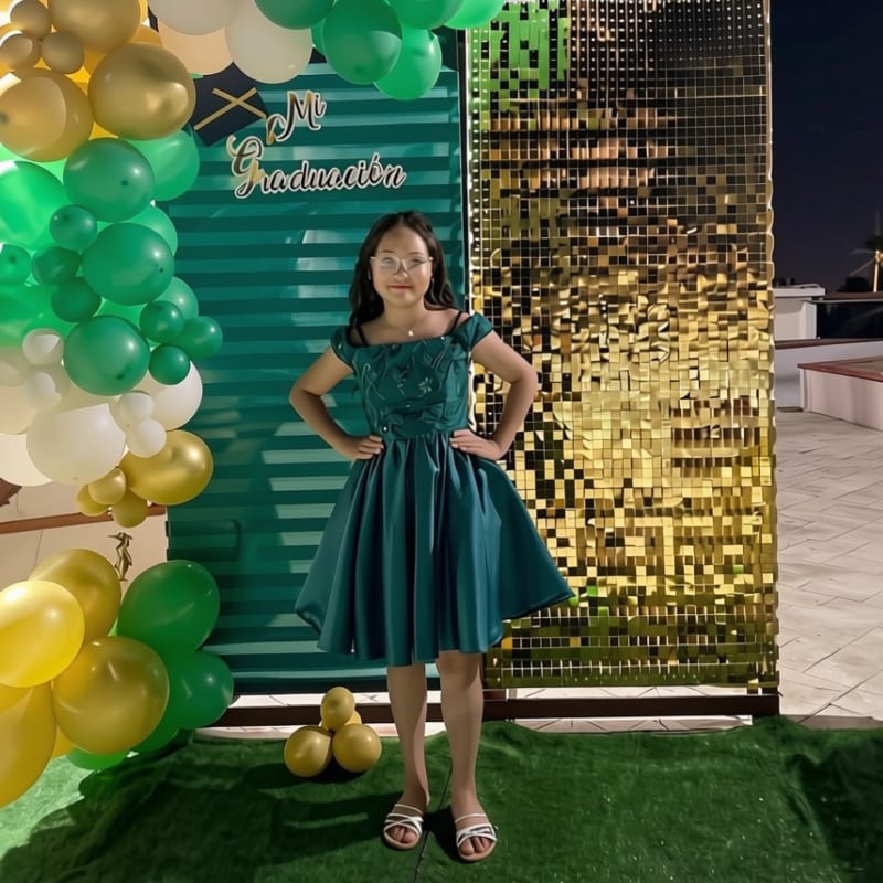 A young girl in a green dress poses in front of a backdrop of green and gold balloons and a shimmering gold sequin wall.