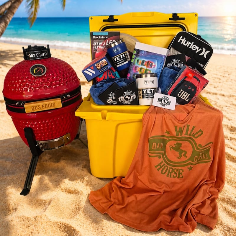 A red ceramic grill and a yellow cooler filled with various items sit on a sandy beach with the ocean in the background.