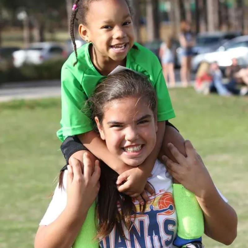 Two smiling girls, one on the shoulders of the other, are in the foreground with a blurred park scene in the background.