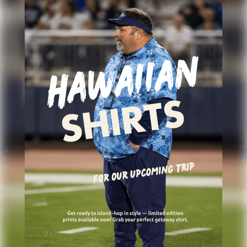 A man in a blue Hawaiian shirt stands on a football field with stadium seating in the background.