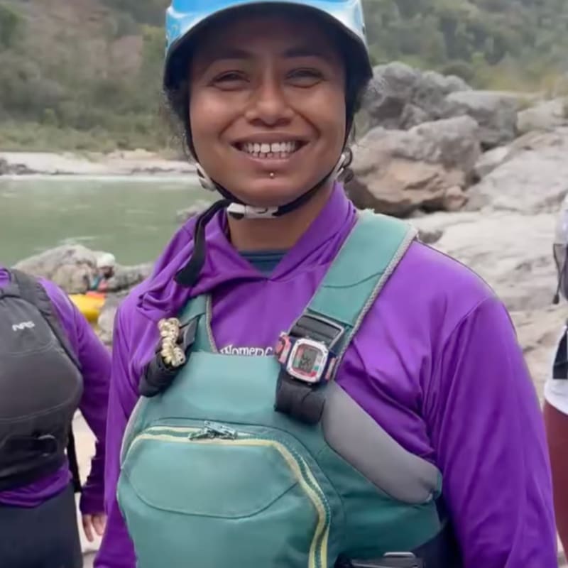 A smiling woman in a purple hoodie and life vest stands in the foreground with a rocky riverbank and calm water in the background.