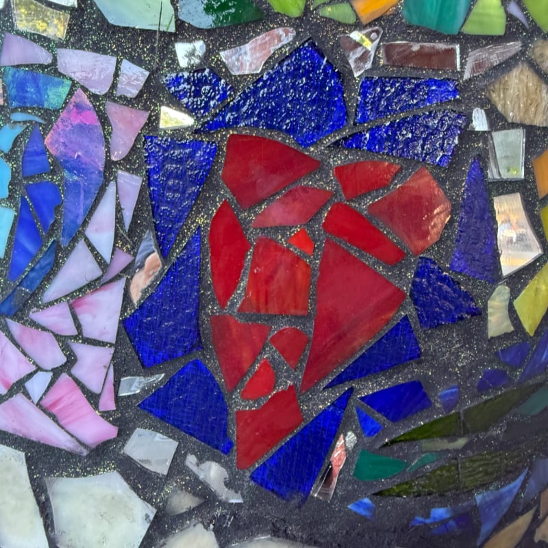 A vibrant mosaic of colorful glass shards, predominantly red and blue, forms a heart shape in the foreground against a dark, textured background.