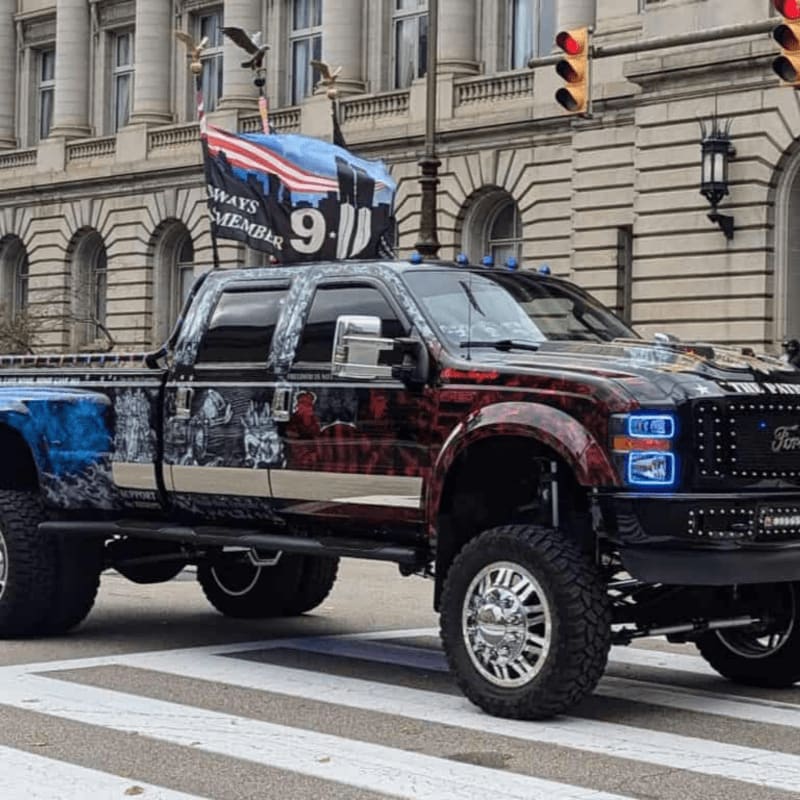 A custom-painted pickup truck with a patriotic theme, featuring the American flag and "9/11 Always Remember" text, is parked on a crosswalk in front of a grand building.