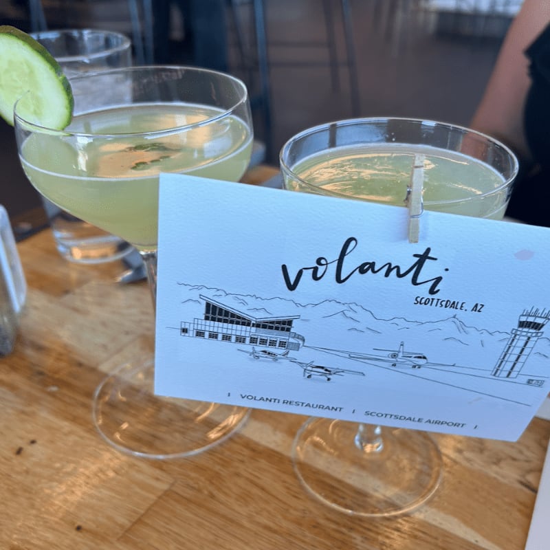Two cocktails with cucumber garnishes sit on a wooden table in front of a card advertising Volanti Restaurant at Scottsdale Airport, with a blurred background of a restaurant interior.