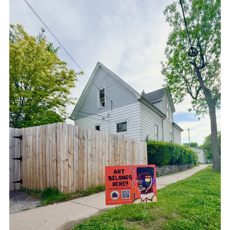 A bright orange sign with "ART BELONGS HERE!!" printed on it stands on a sidewalk in front of a wooden fence and a two-story house with a cloudy sky above.