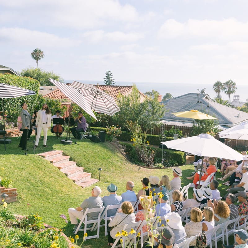 A wedding ceremony is taking place outdoors on a sunny day, with guests seated on a lawn in the foreground and a band performing in the background, overlooking the ocean.