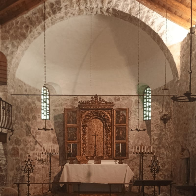 A rustic altar with a wooden altarpiece stands in the foreground, while the background reveals a stone-walled church interior with arched ceilings and windows.