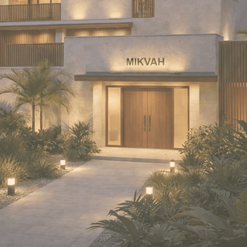 A well-lit pathway leads to the "MIKVAH" entrance of a modern building, surrounded by lush tropical foliage.