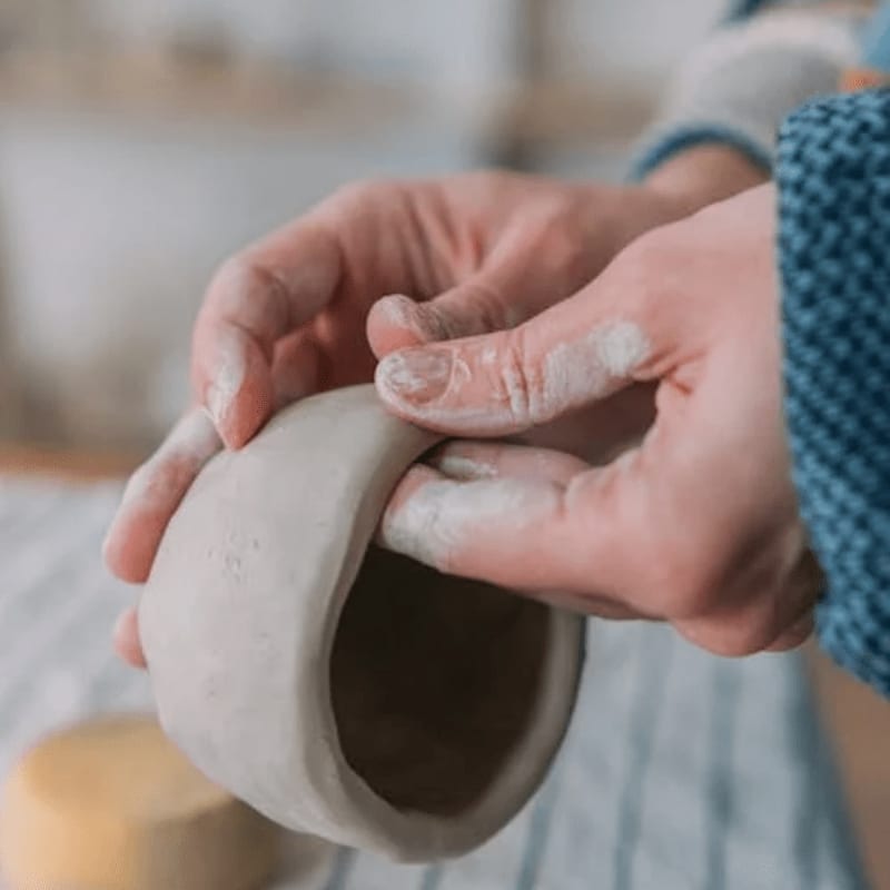 Hands shape a clay pot in the foreground, with a blurred background of a workshop.