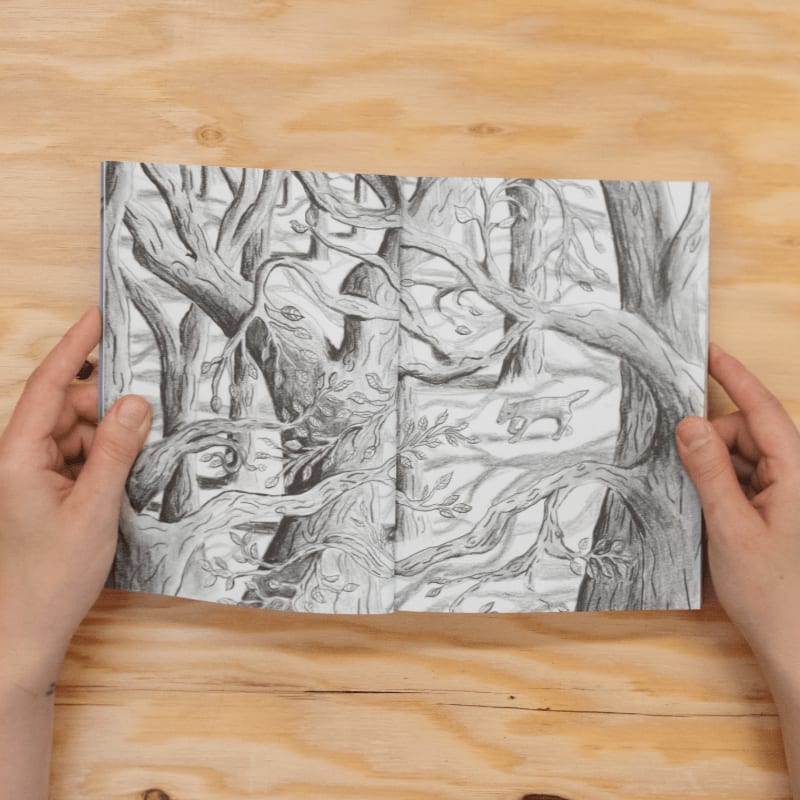 Hands hold open a sketchbook filled with a detailed pencil drawing of a dense forest with a tiger lurking in the background.
