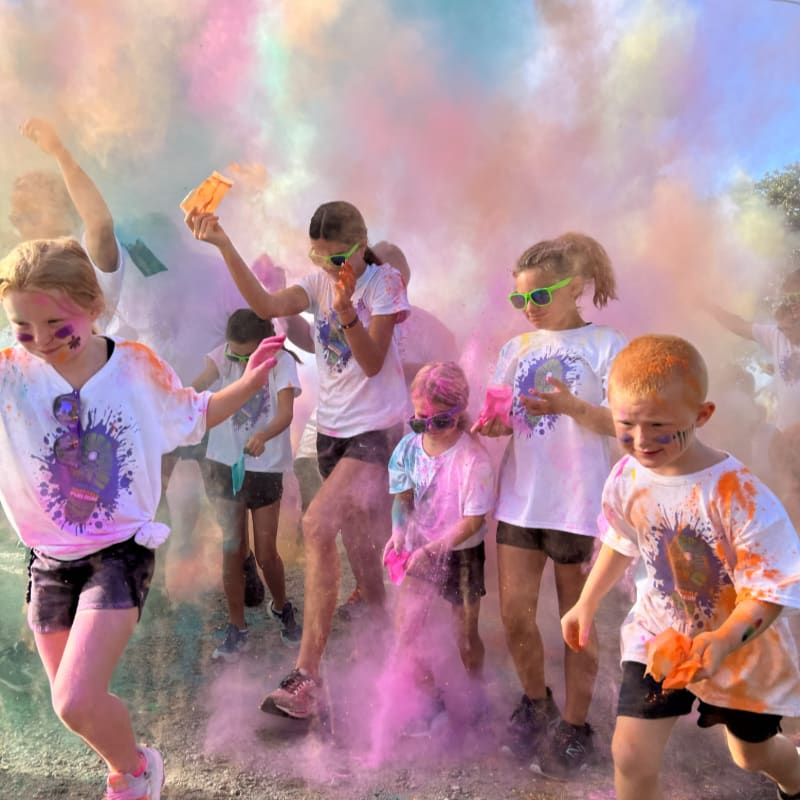 Children in white shirts run through clouds of colorful powder, with a bright sky and trees visible in the background.