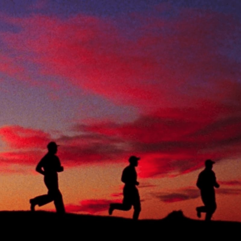Three silhouetted figures run across a grassy hill against a vibrant sunset sky.
