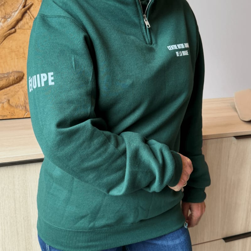 A person in a green quarter-zip sweatshirt with "ÉQUIPE" on the sleeve stands in front of a wooden dresser and a framed artwork.