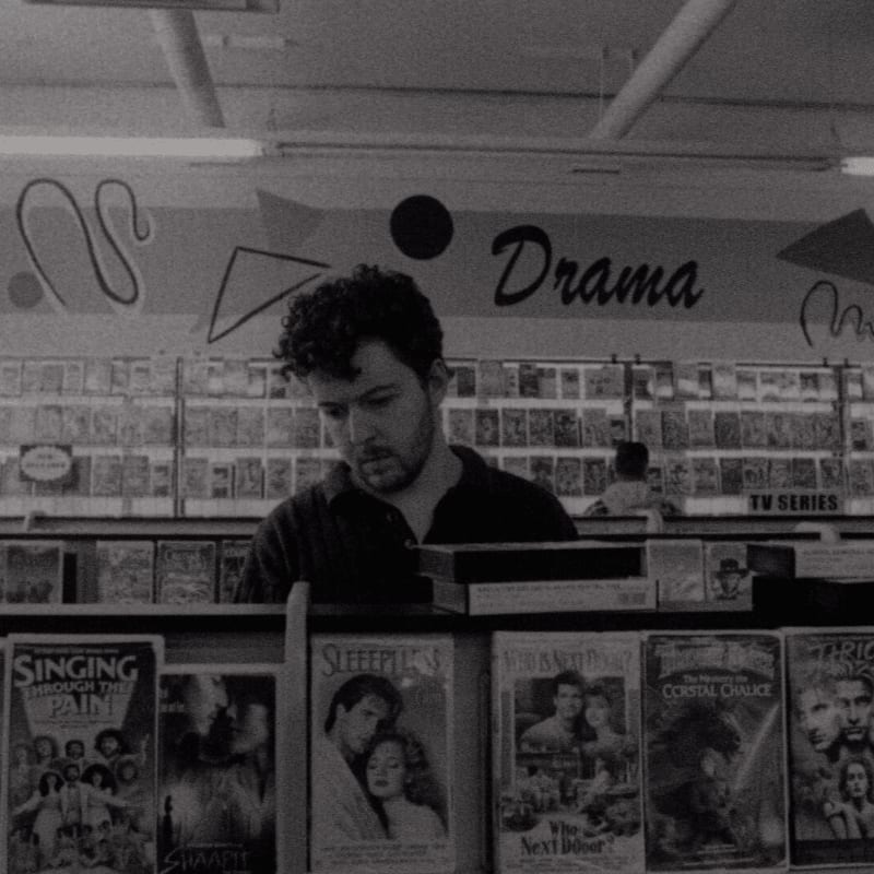 A man browses a shelf of DVDs in a store with "Drama" and "TV Series" signs visible in the background.