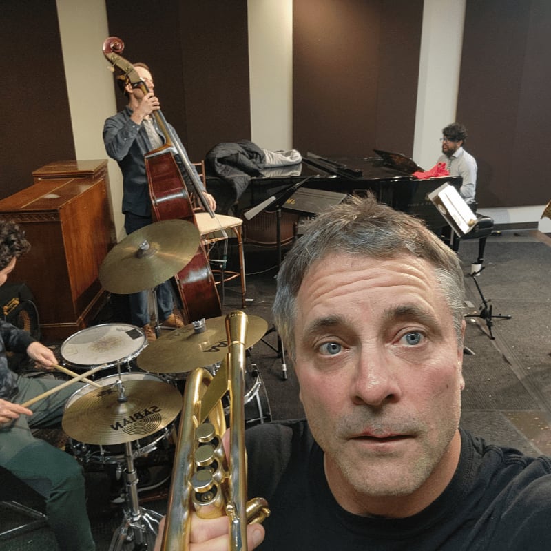 A man holds a trumpet in the foreground, while in the background, a band plays jazz with a double bass, drums, and piano.