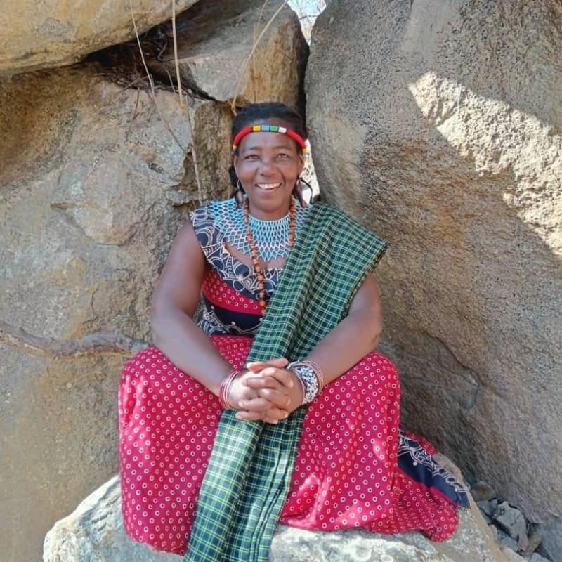 A smiling woman in traditional attire sits between large rocks, with a dry, rocky landscape in the background.