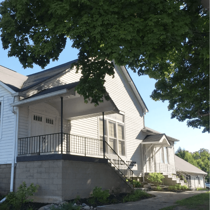 A white building with a porch and stairs is framed by the green leaves of a tree in the foreground, with a clear blue sky in the background.
