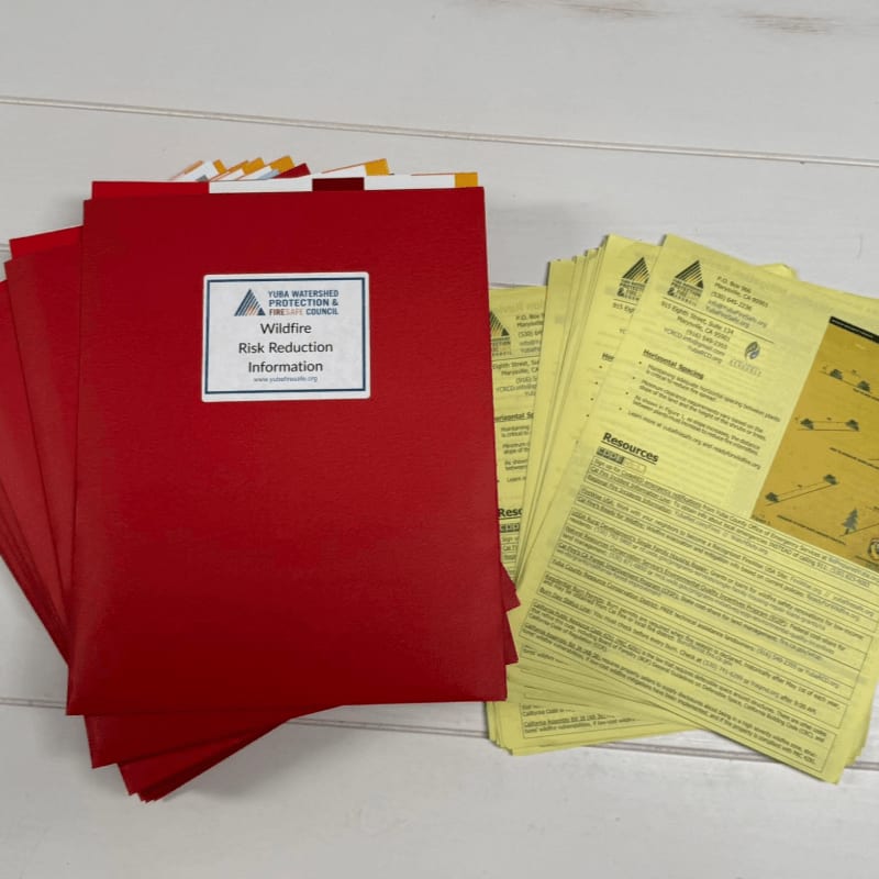 A stack of red folders labeled "Wildfire Risk Reduction Information" sits in the foreground next to a pile of yellow informational sheets, all on a light-colored wooden surface.