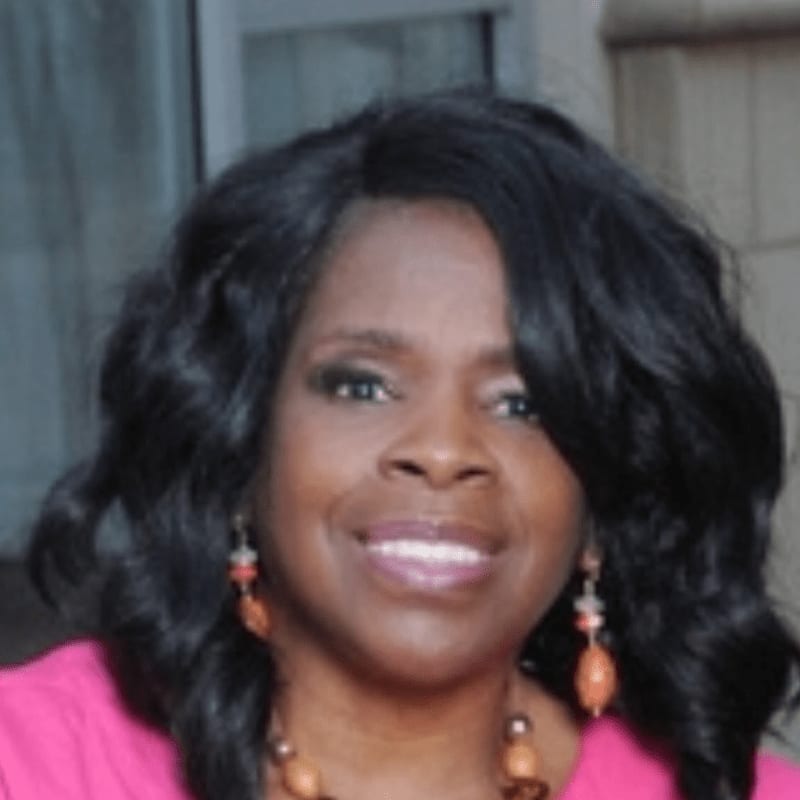 A smiling Black woman with dark, wavy hair and a pink top is in the foreground, with a blurred background of what appears to be an outdoor setting.
